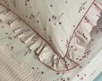 Può includere: Una federa e un lenzuolo rosa chiaro abbinati con un delicato motivo floreale rosa scuro. La federa è rifinita con un bordo a volant e un sottile profilo rosa scuro. Una coperta in maglia rosa chiaro è posizionata sopra il lenzuolo.