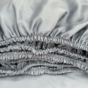 May include: A light gray fitted sheet featuring a smooth, satin-like fabric texture. The edges of the sheet are gathered with elastic, creating a series of soft, uniform ruffles and folds that extend across the width of the textile.