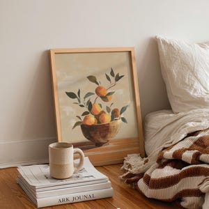 May include: A framed print of oranges in a bowl with green leaves. A beige mug sits on a stack of books with the text "ARK JOUNAL". The image is on a wooden floor next to a bed with a striped blanket.