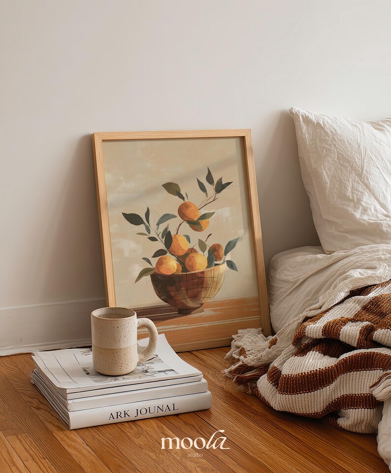 May include: A framed print of oranges in a bowl with green leaves. A beige mug sits on a stack of books with the text "ARK JOUNAL". The image is on a wooden floor next to a bed with a striped blanket.