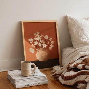May include: A framed art print featuring a vase of white flowers against a burnt orange background. A ceramic mug sits on a stack of books, with a striped blanket and pillow in the background. The print is in a light wood frame.