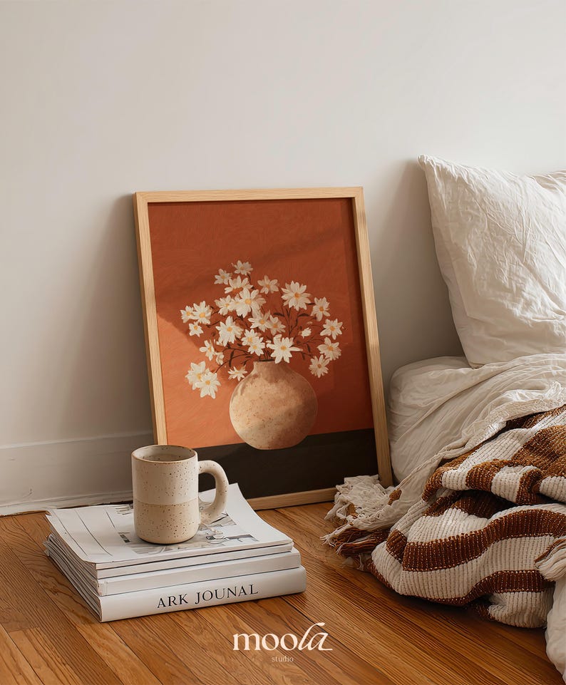 May include: A framed art print featuring a vase of white flowers against a burnt orange background. A ceramic mug sits on a stack of books, with a striped blanket and pillow in the background. The print is in a light wood frame.