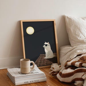 May include: A framed art print featuring a white dog wearing sunglasses, sitting on a rooftop under a full moon. A beige mug rests on a stack of books, with the text "ARK JOUNAL" visible. The artwork is set against a dark background.