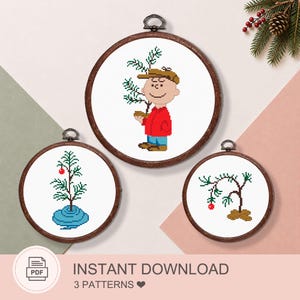 May include: Three circular wooden embroidery hoops containing cross-stitch patterns. One features a cartoon character in a red shirt holding a small tree, another shows a small tree with a red ornament, and the third depicts a sparse tree with one ornament.