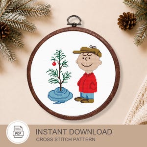 May include: A circular wooden embroidery hoop containing a cross stitch pattern of a cartoon character wearing a tan cap, red jacket, and blue pants standing next to a small green Christmas tree with a single red ornament on a blue base.