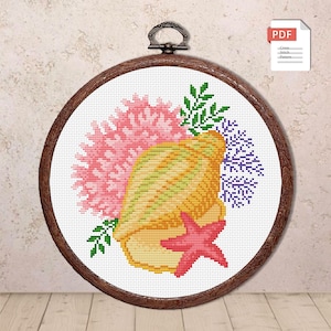 May include: A cross-stitch pattern of a seashell, starfish, and coral in a wooden embroidery hoop. The seashell is yellow and green, the starfish is red, and the coral is pink. Green leaves and purple accents complete the design.