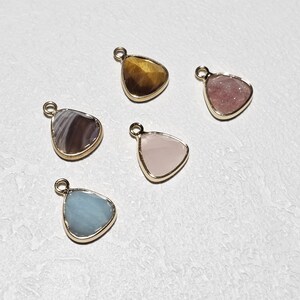 Puede incluir: Cinco colgantes de piedras preciosas con borde de metal dorado. Las piedras preciosas son en tonos marrón, rosa, azul y gris. Cada colgante tiene un pequeño lazo en la parte superior para sujetar.