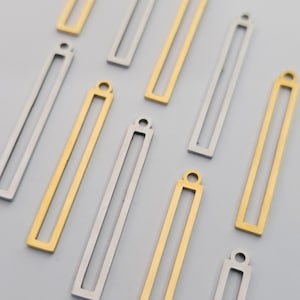 May include: A collection of rectangular metal charms in gold and silver tones. Each charm features a long, thin, open-frame design with a small circular loop at the top for attachment. The pieces are arranged in a scattered pattern on a neutral surface.