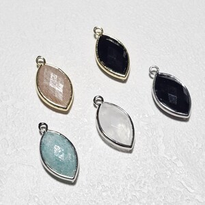 Puede incluir: Una colección de colgantes de piedras preciosas en forma de marquesa. Los colgantes presentan varios colores, incluyendo negro, rosa claro, blanco y azul-verdoso, cada uno engastado en un marco de metal dorado o plateado. Las piedras tienen un corte facetado.