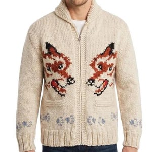 May include: A cream-colored knit zip-up cardigan featuring a shawl collar and two front pockets. The sweater displays two large, symmetrical fox head patterns in shades of brown, black, and white, with small paw print motifs along the bottom hem.