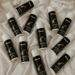 May include: A collection of black and white spray bottles, each labelled "Pause". The bottles have white caps and a graphic of a hand holding a stopwatch. They are arranged on a white, textured surface, creating a visually appealing display.
