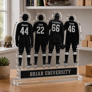 Op de afbeelding: Een heldere acryl display met vier hockeyspelers in zwarte shirts met witte nummers en namen. De basis leest "BRIAR UNIVERSITY" in witte tekst op een zwarte achtergrond. De spelers dragen hockeyschaatsen.