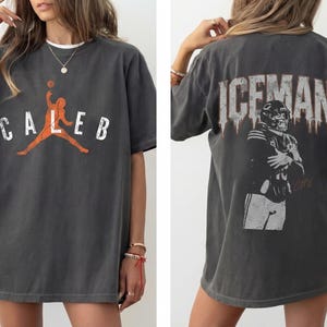 May include: Dark gray oversized t-shirt featuring the name "CALEB" with a basketball player graphic in orange. The back of the shirt displays the word "ICEMAN" above a football player illustration. A casual, sporty apparel item.