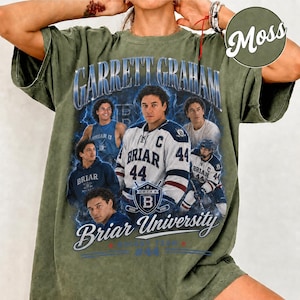 May include: Olive green t-shirt featuring a graphic of a hockey player with the text "Garrett Graham" and "Briar University." The design includes multiple images of the player in various poses, with the number 44 prominently displayed on the jersey.