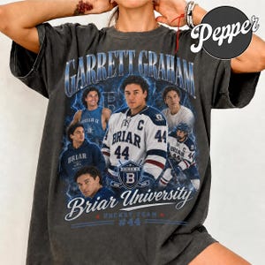 May include: Dark gray t-shirt featuring a graphic of Garrett Graham and the Briar University hockey team. The design includes multiple images of a hockey player, the text "Briar University Hockey Team #44", and lightning bolt accents.