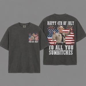 以下が含まれることがあります： チャコールグレーのTシャツで、保安官の制服とカウボーイハットを身に着けた男性が花火の上がるアメリカ国旗の前に立っているグラフィックが特徴です。背面には「HAPPY 4TH OF JULY TO ALL YOU SUMBITCHES」という文字がプリントされ、前面にはその縮小版が配置されています。
