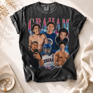 Può includere: T-shirt grigio scuro con un collage di immagini e il nome "GRAHAM" in un carattere retrò colorato. La maglietta presenta immagini di diverse persone, con il nome "Briar" e stelle sotto.