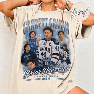 May include: Cream-colored t-shirt featuring a graphic of a hockey player with the text "Garrett Graham" and "Briar University Hockey Team #44". The design includes multiple images of the player in various poses, with a vintage aesthetic.