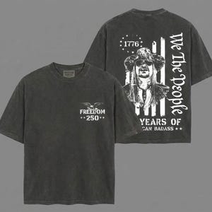 May include: Dark grey t-shirts with white graphics. One shirt features an eagle and the text "FREEDOM **250**". The other shirt has a portrait, the text "We The People", "1776", and "YEARS OF CAN BADASS **". The shirts are part of the American Heritage collection.