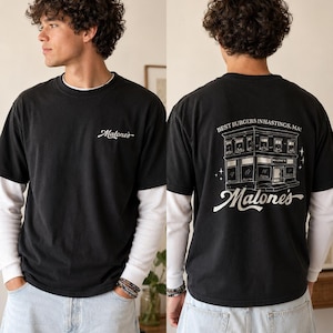 以下が含まれることがあります： 前面に白い文字で「Malone's」と書かれた黒いTシャツ。背中には建物の詳細なイラストと「Best Burgers in Hastings, MA!」と「Malone's」の文字が描かれています。