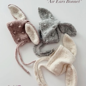 May include: Three knitted bonnets labeled Air Ears Bonnet, each featuring long, upright ears and small bobble details. The bonnets are in dusty rose, heather gray, and cream, all crafted with chin ties for securing the head covering in place.