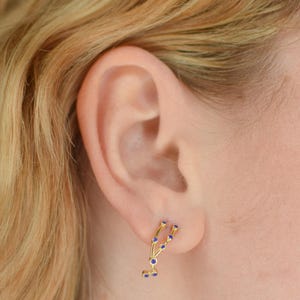 May include: Gold-colored earring with a constellation design, featuring small blue gemstones. The earring is worn on a person's ear, with blonde hair visible in the background. The jewelry is a delicate and stylish accessory.