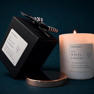 May include: A lit candle in a white glass jar with a label reading The White Garden, next to a black gift box with a black and white striped ribbon and a metallic copper lid. The label specifies 100% rapeseed & coco wax and origin.