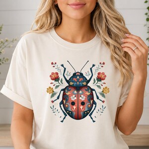 May include: A cream-colored t-shirt featuring a central graphic of a ladybug adorned with intricate blue, red, and white floral patterns. The ladybug is surrounded by small, delicate botanical illustrations of red and yellow flowers and green leaves.