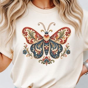 May include: A cream-colored short-sleeve t-shirt featuring a large, centered graphic of a butterfly. The butterfly is decorated with intricate, symmetrical floral and scroll patterns in shades of deep red, navy blue, and muted gold.