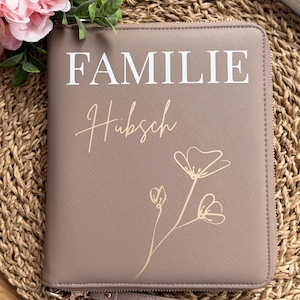 Könnte beinhalten: Eine taupefarbene Reißverschluss-Geldbörse mit dem Wort "FAMILIE" in weißen Blockbuchstaben und "Hübsch" in Schreibschrift. Ein goldenes Blumendesign befindet sich unten rechts. Die Geldbörse hat eine Handschlaufe und liegt auf einer gewebten Matte.