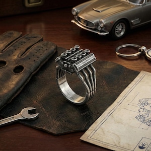 May include: A silver-toned ring shaped like a V8 engine block with the word MASERATI engraved on the side. The ring features detailed exhaust pipes extending down the band, mimicking the mechanical structure of an automotive engine component.