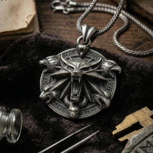 May include: A silver-colored pendant necklace featuring a detailed circular design with a wolf's head at the center, surrounded by animal figures and inscriptions. The pendant hangs from a braided chain. The image also shows a book and other tools.