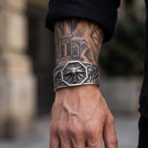 May include: A wide silver-toned metal cuff bracelet featuring a central octagonal emblem with a detailed wolf head relief. The surface of the cuff has a textured, rugged finish surrounding the central design, creating a bold and industrial appearance.