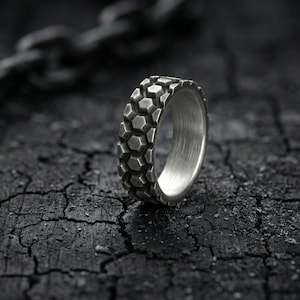 May include: A silver ring with a tire tread design. The ring is made of metal and has a textured surface. The ring is set against a dark, textured background with a chain in the background.