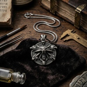 May include: A silver pendant necklace featuring a wolf's head design with intricate details. The pendant is on a silver chain, resting on a dark cloth. Other items include a book, a brush, and a small bottle.