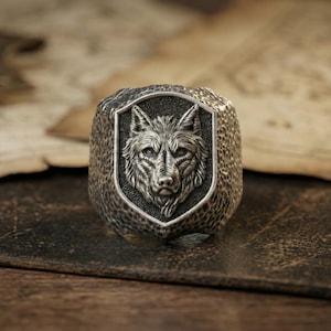 May include: A silver-toned ring featuring a detailed wolf head design within a shield-shaped frame. The ring has a textured surface and is set against a backdrop of aged paper and leather. The ring is a piece of jewelry.