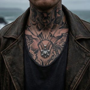 May include: A silver pendant necklace with a detailed design hangs over a black shirt. The person is wearing a brown leather jacket. The person has tattoos on their chest and neck. The background is a blurred view of the ocean.