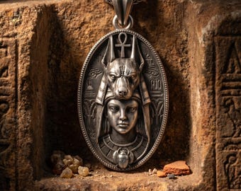 May include: An oval silver pendant featuring a relief of a human face topped with an Anubis head, an ankh symbol, and a scarab beetle at the base. The pendant is suspended from a silver chain and displays intricate Egyptian-style hieroglyphic carvings.