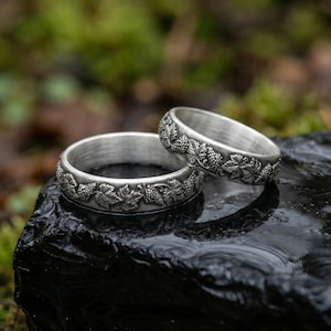 Könnte beinhalten: Zwei silberne Ringe mit detailliertem Trauben- und Blattdesign. Die Ringe werden auf einer dunklen, nassen Oberfläche mit einem verschwommenen grünen und braunen Hintergrund präsentiert. Die Ringe haben ein strukturiertes, graviertes Muster.
