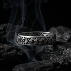 May include: A silver ring with an intricate geometric pattern, resting on dark charcoal. Wisps of smoke rise around the ring, creating a dramatic effect. The ring's design is a repeating series of interlocking shapes.
