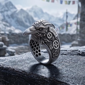 May include: A silver-toned metal ring featuring a detailed raven head design integrated with mechanical gear and piston motifs. The inner band of the ring displays a hexagonal honeycomb pattern, while the exterior showcases intricate metallic textures.