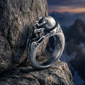 Puede incluir: Un anillo de plata con una figura esculpida abrazando un globo terráqueo. El diseño del anillo incluye una representación detallada de una persona sosteniendo la Tierra, sobre un fondo rocoso. El anillo es de metal.