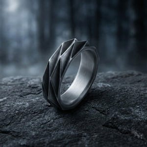 May include: A silver ring with a geometric design. The ring features a series of angled, triangular facets that wrap around the band. The ring is set against a dark, textured stone, with a blurred forest background.