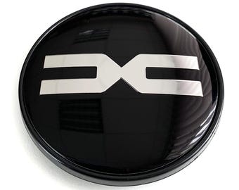 May include: A glossy black car emblem with a silver logo. The emblem is circular with a raised edge. The silver logo is a stylized design of the letters "DC".