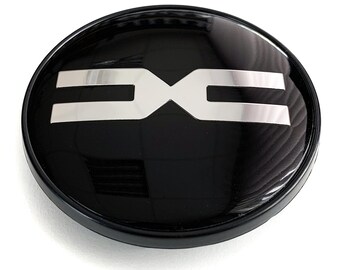May include: A black, glossy, circular emblem with a silver, stylized "DX" logo. The emblem has a raised, rounded edge and a reflective surface, creating a modern and sleek appearance. The logo is centered on the emblem.