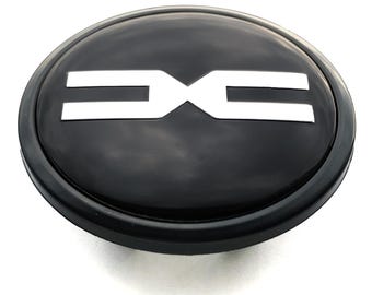 May include: A black, oval-shaped car emblem with a white logo. The logo consists of two horizontal lines connected by a vertical line in the center. The emblem has a glossy finish.
