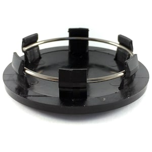Peut inclure: Un cache-moyeu circulaire en plastique noir doté d'une surface supérieure lisse et plate, ainsi que de six clips verticaux sur la face inférieure. Un fin ressort de tension métallique argenté est inséré dans les clips pour fixer le cache.