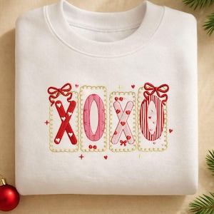 May include: A white sweatshirt featuring an embroidered "XOXO" design in red and pink. The letters are framed in gold with heart and bow details. A red bauble and a pine branch are also visible.