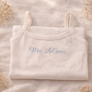 May include: Cream-colored tank top with thin straps, folded neatly. The text "Mrs. LaCascia" is embroidered in light blue. The top is set on a soft, light-colored fabric, with dried flowers in the background.