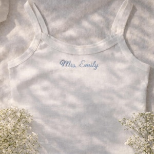 Könnte beinhalten: Weißes Tanktop mit dünnen Trägern, bestickt mit dem Text "Mrs. Emily" in Hellblau. Das Oberteil ist aus einem weichen Stoff gefertigt und wird auf einer strukturierten Oberfläche präsentiert, mit weißen Blumen am unteren Rand.